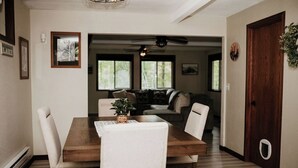 Ein Essbereich mit Holztisch und Stühlen, einem Teppich und einer Pflanze auf dem Tisch. Eine Tür befindet sich rechts, und ein Flur führt in ein Wohnzimmer mit einer Couch und einem Deckenventilator.
