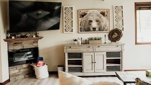 Ein Wohnzimmer mit Kamin aus Stein, einem an der Wand montierten Fernseher, einem weißen Schrank mit Dekoration und einem großen Bärenbild.