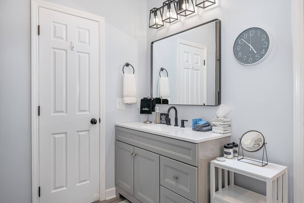 A bathroom with a white door, a vanity with a mirror, a sink, and a clock on the wall.
