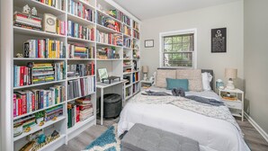 A bedroom with a large bookshelf, a bed with a grey and white duvet, a window with blinds, and a wall-mounted lamp.