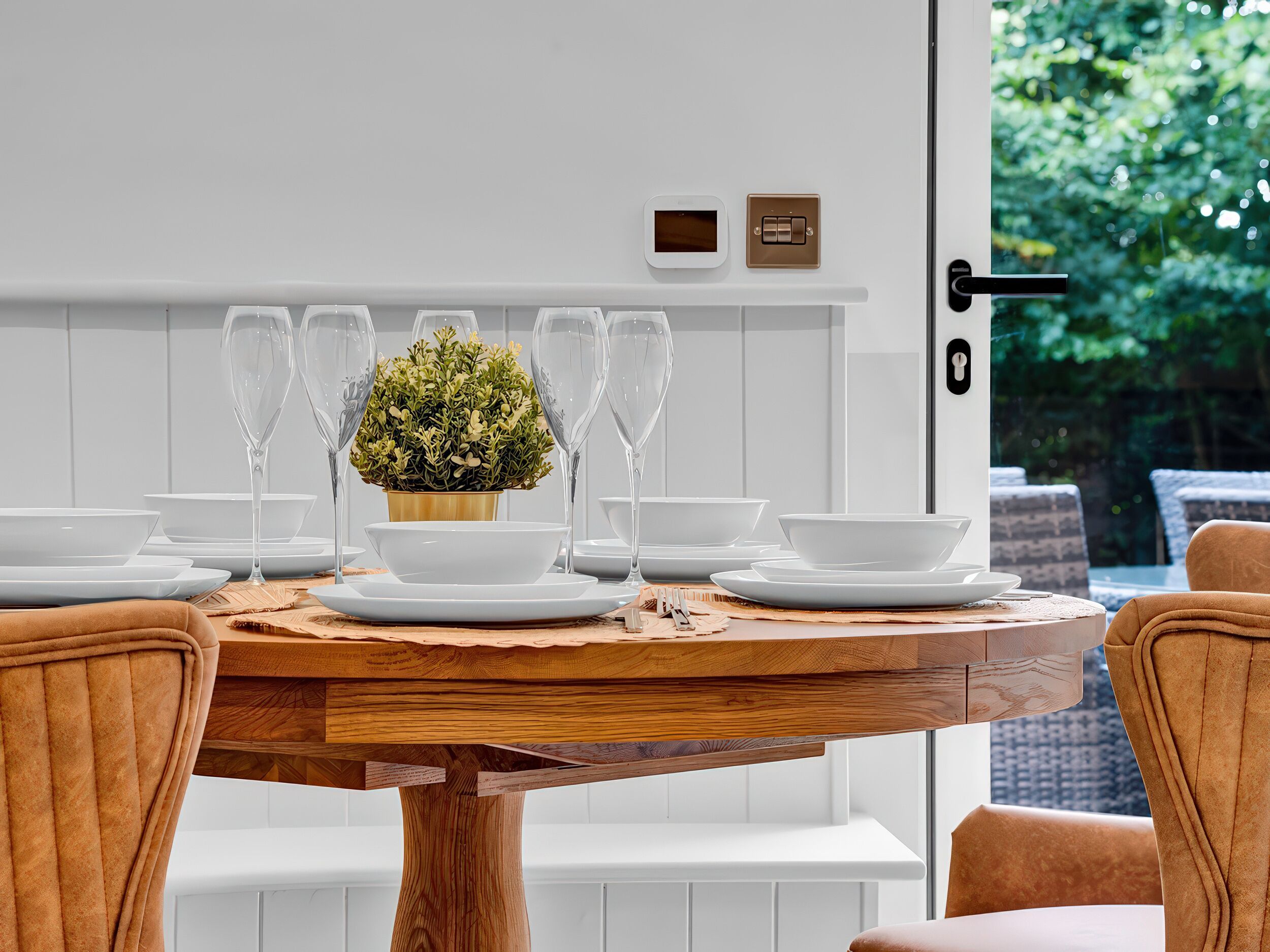 Una mesa de comedor dispuesta con platos, tazones y vasos blancos, adornada con una planta en maceta en el centro.