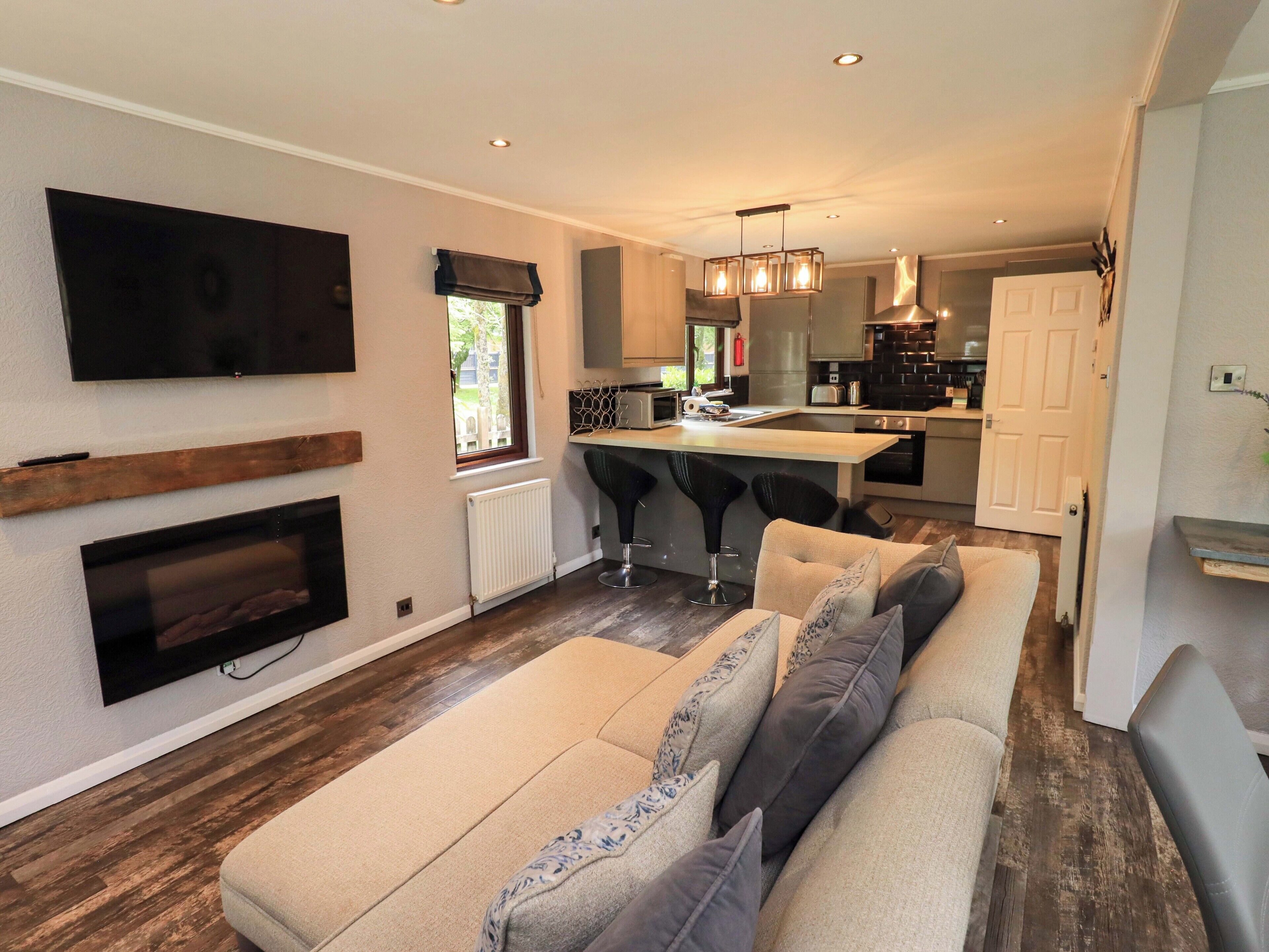 A modern living room with a large flat-screen TV, a fireplace, a sofa with patterned cushions, and a kitchen area with a bar stool.