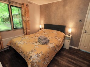 Ein Schlafzimmer mit Blumenmuster-Bettdecke, einem ordentlich bezogenen Bett mit gefalteten Handtüchern, einem Nachttisch mit Lampe und einem Fenster mit Vorhängen.