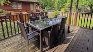 Une terrasse extérieure couverte, dotée d'une table à plateau de verre et de chaises en osier, entourée d'une terrasse en bois et d'une rambarde en métal.