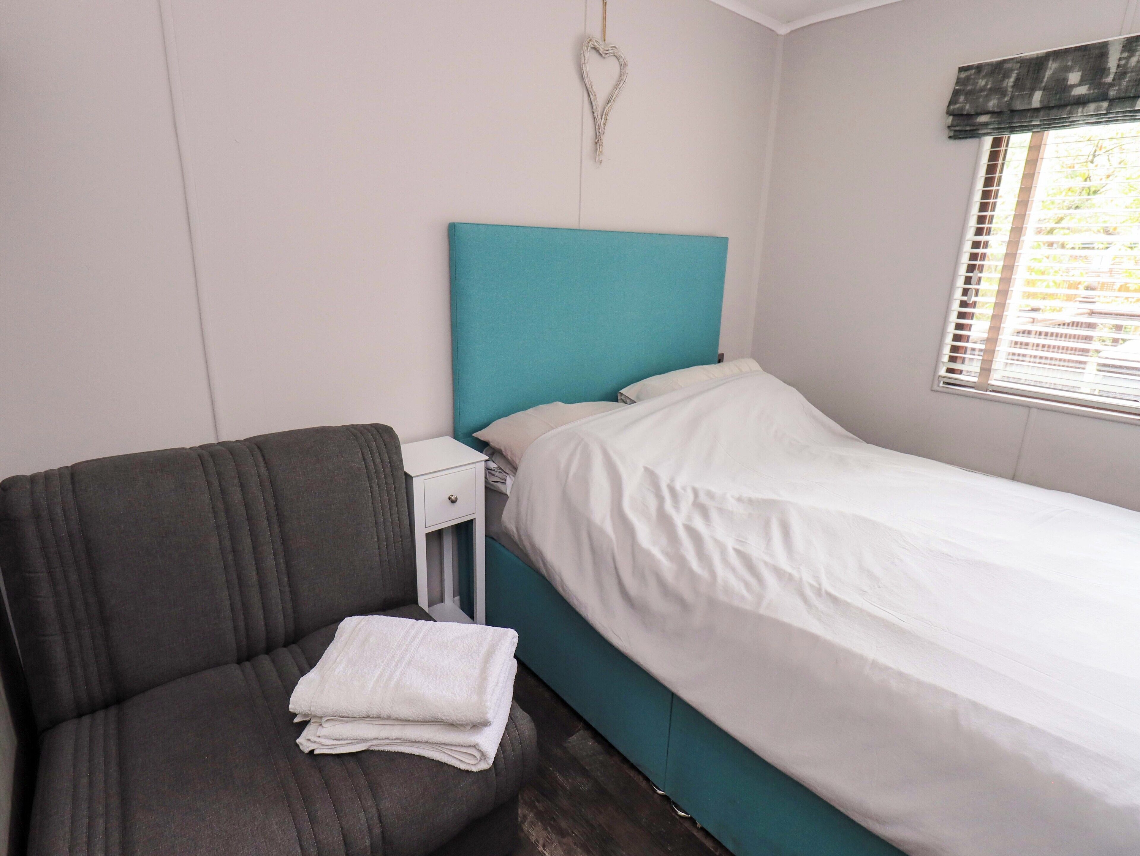A single bed with a white mattress and teal headboard, a small bedside table with a drawer, and a grey upholstered chair.