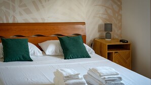 Ein ordentlich bezogenes Bett mit weißen Leinentüchern und zwei grünen Kissen, ein Nachttisch mit Lampe und Telefon sowie ein Holzbeistelltisch mit einer kleinen Ablagefläche.