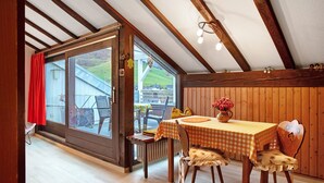 Un coin repas avec une table dressée pour deux personnes, une chaise à motifs floraux et une vue sur un paysage verdoyant par une porte vitrée.