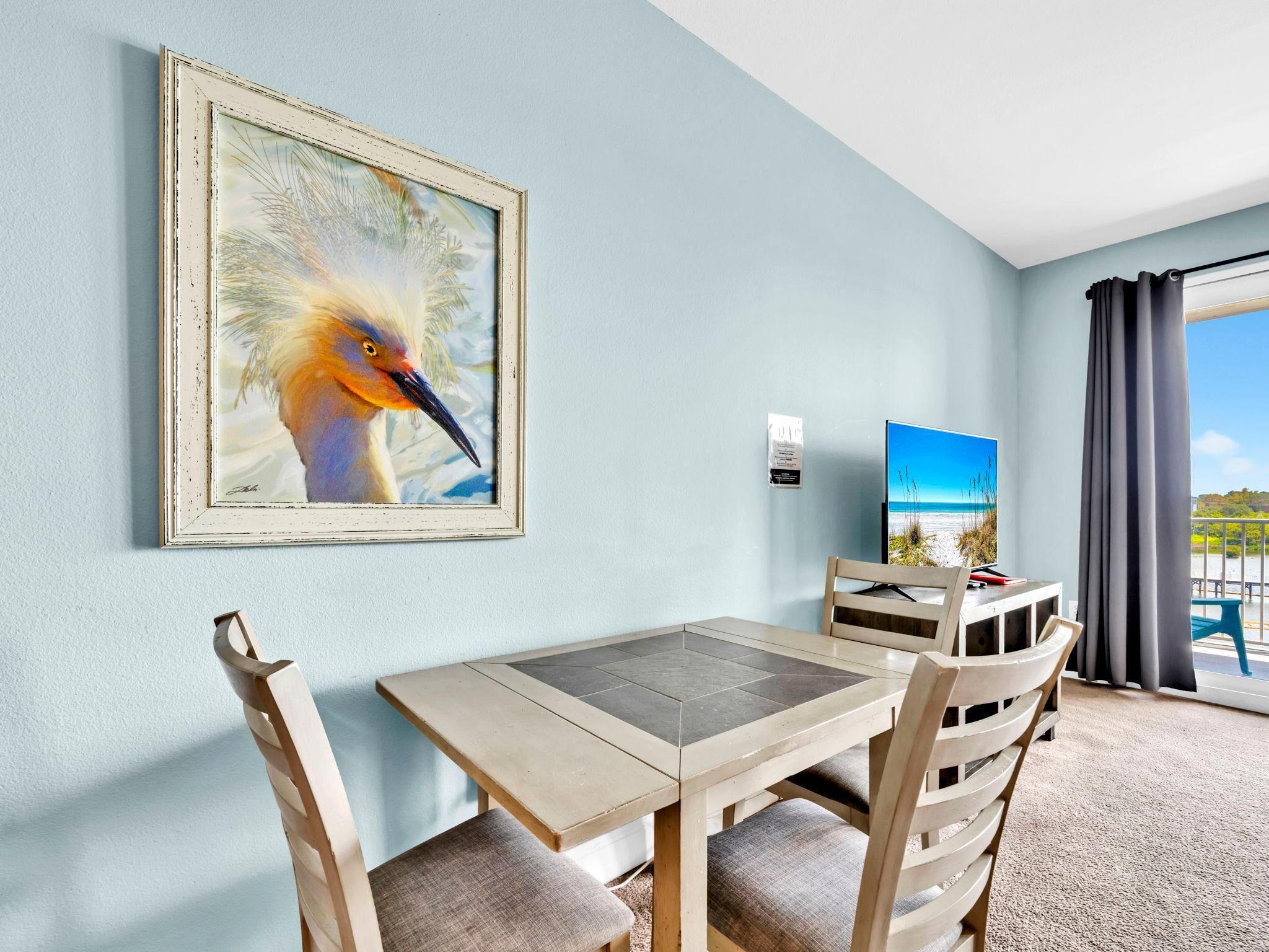 A hotel room with a dining table, chairs, a framed painting of a bird, a television, and a view of the beach.