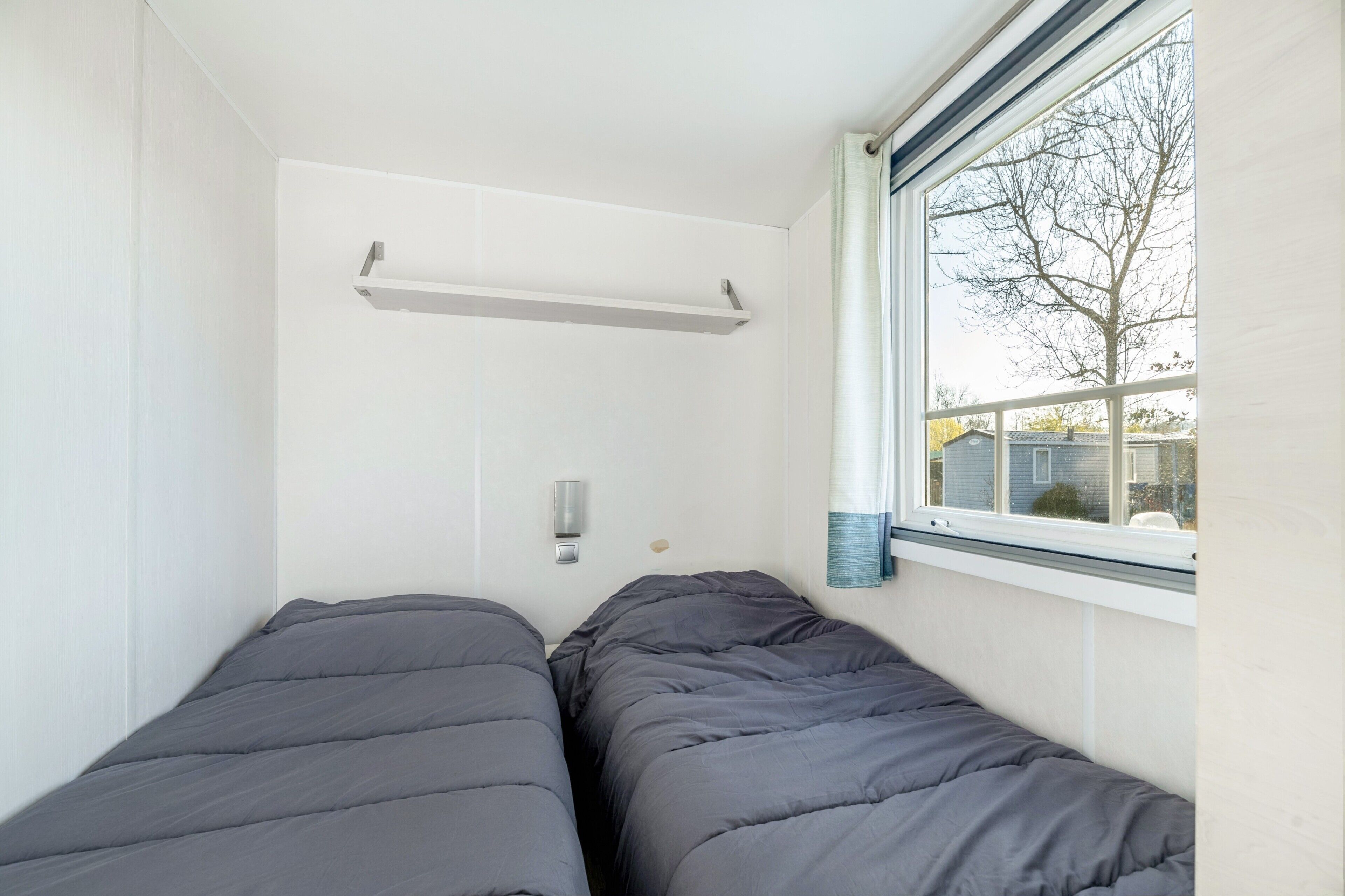 A small, single room with two beds, a window, and a shelf.