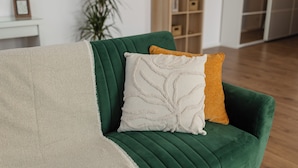 Un sofá verde con una manta color beige y dos almohadas, una blanca y otra mostaza, en una habitación moderna con suelo de madera y una planta al fondo.