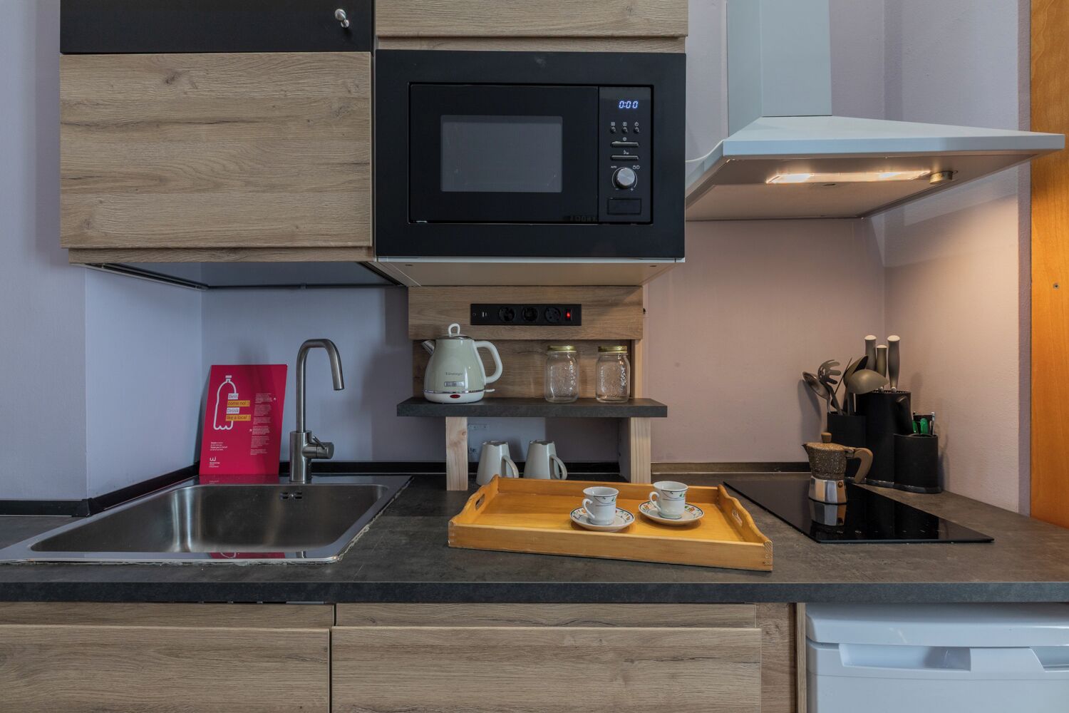 A modern kitchen with a microwave, kettle, and coffee maker on a wooden shelf.