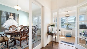 A dining area with a wooden table and chairs, a painting on the wall, and a view through a glass door to a room with a window and a chandelier.
