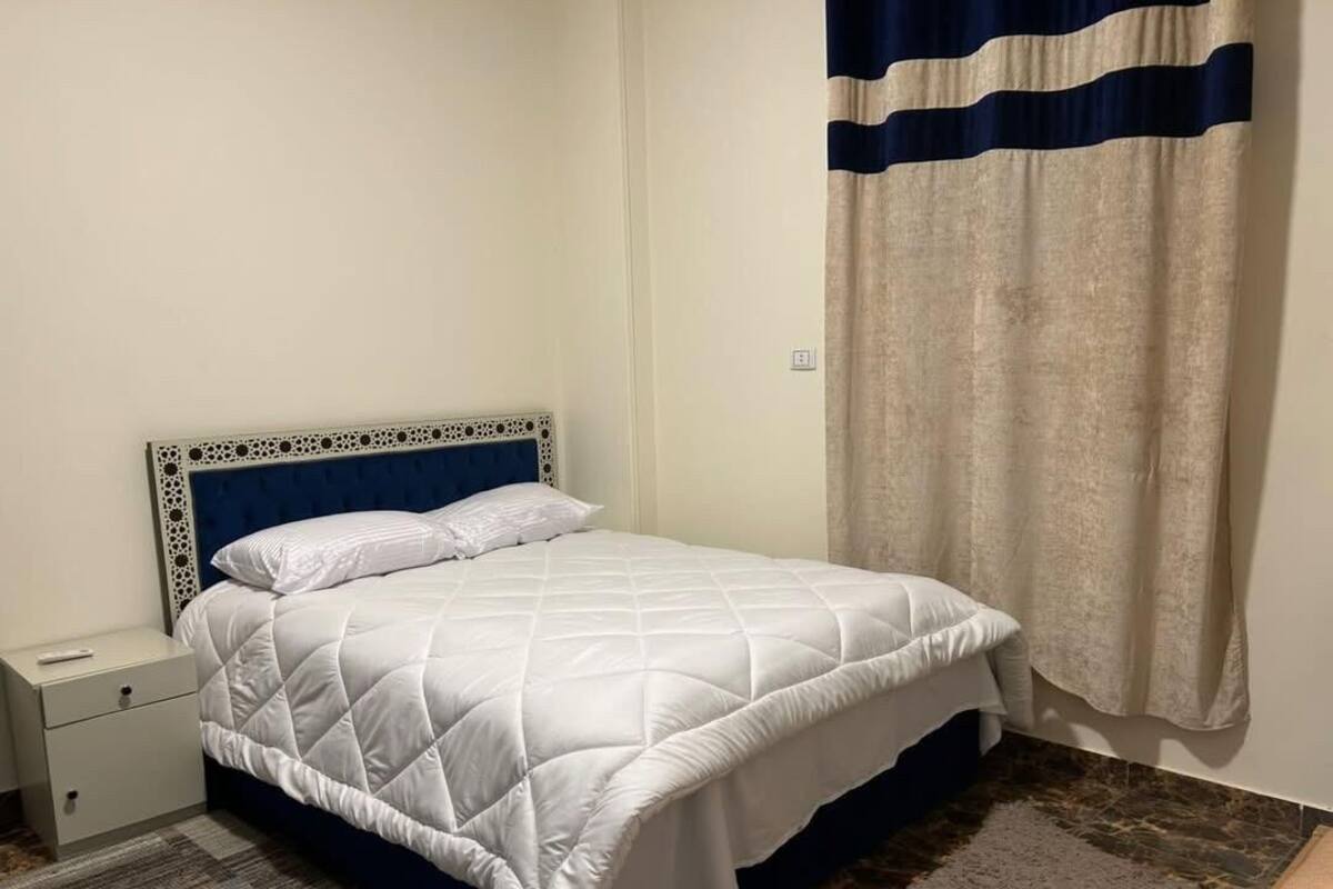 A neatly made bed with a white quilt and a blue headboard, a small bedside table, and a curtain with horizontal stripes.