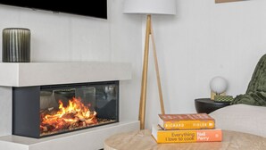 A modern living room with a fireplace, a television mounted on the wall, and a round table with books. - Jamburra - Breathtaking views of the Blue Mountains! (Leura)
