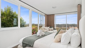 A bedroom with a large bed, multiple pillows, a blanket, and a view of the outdoors through large windows. - Jamburra - Breathtaking views of the Blue Mountains! (Leura)