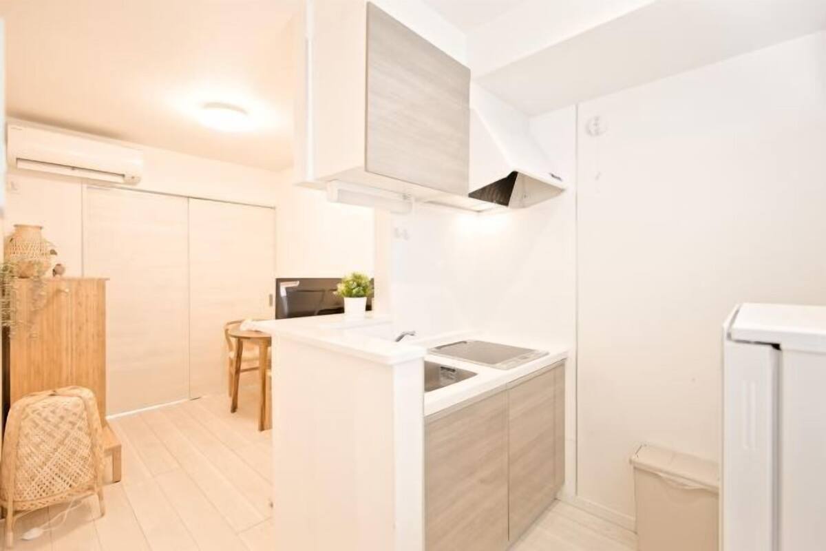 Cocina compacta con encimera blanca, un fregadero y horno empotrado. Hay una silla de madera y una mesa pequeña con una planta.