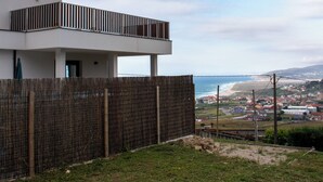 Un edificio moderno con techo plano y balcón, con vistas a una zona costera que incluye una playa y casas al fondo.