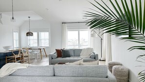 Uma sala de estar moderna com um sofá cinzento, uma mesa de jantar com cadeiras e uma janela grande.