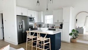 Uma cozinha moderna com armários brancos, uma geladeira de aço inoxidável, uma ilha central com banquetas e uma área de jantar com um sofá cinza.