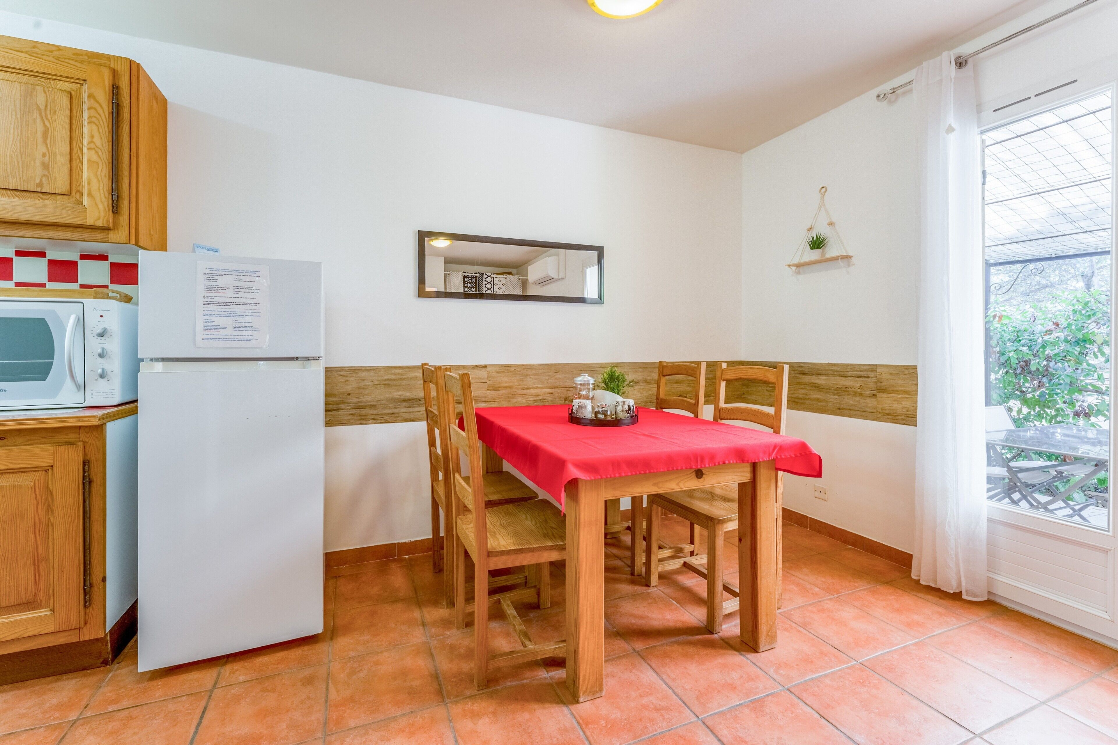 Une cuisine compacte équipée d’un four à micro-ondes, d’un réfrigérateur, de meubles en bois, d’une table rouge avec des chaises, d’un miroir et d’une fenêtre donnant sur un espace verdoyant.