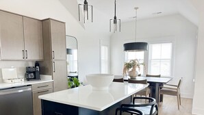 A modern kitchen with a central island, stainless steel appliances, and a dining area with a large window. - Cocoa Beach! Luxury Pet Friendly Beach Home! (Kill Devil Hills)