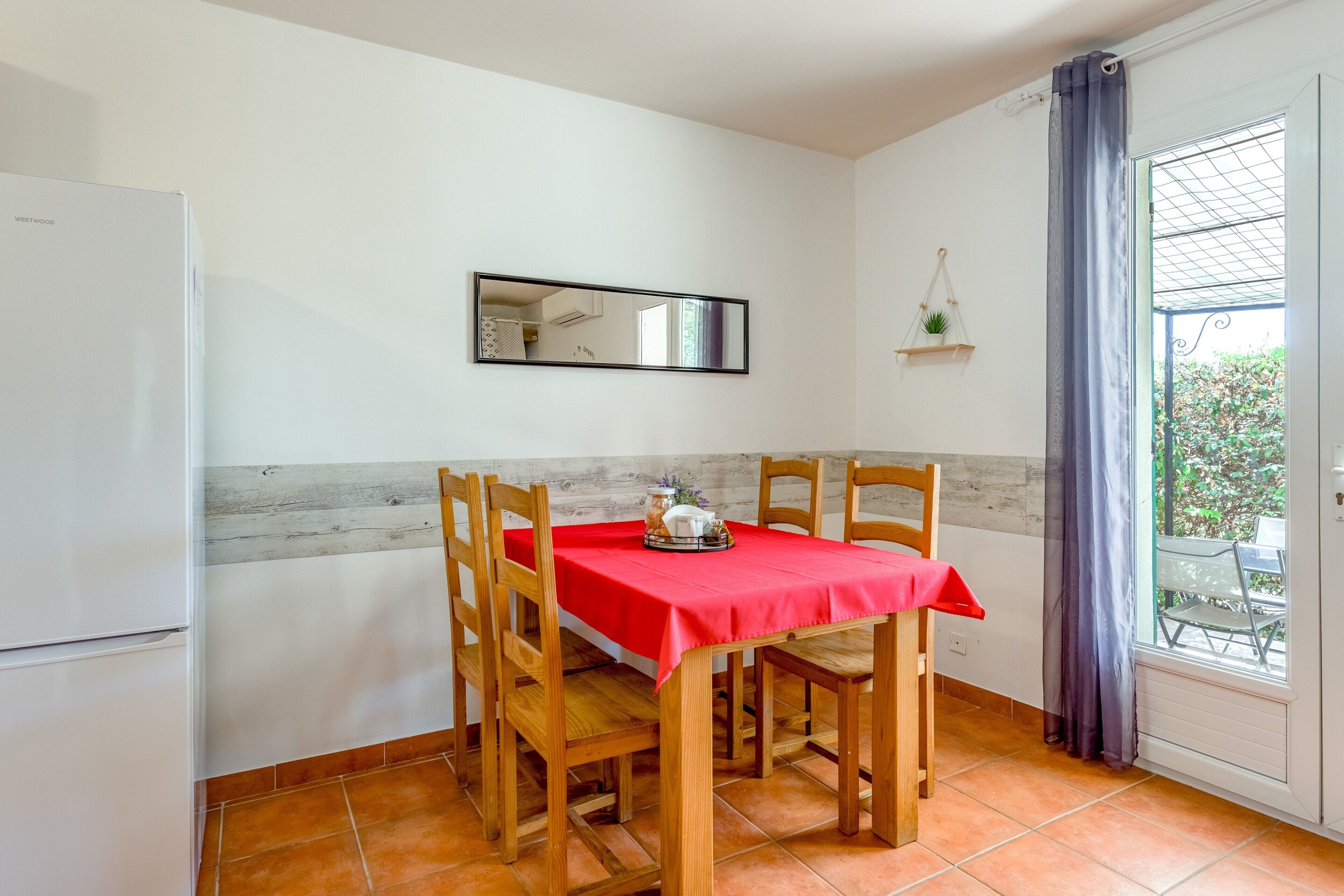 A dining area with a table set for two, wooden chairs, a red tablecloth, a white refrigerator, a mirror, and a window with a view of greenery.