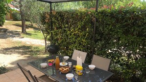 An outdoor patio with a table set for dining, surrounded by greenery and a barbecue grill.