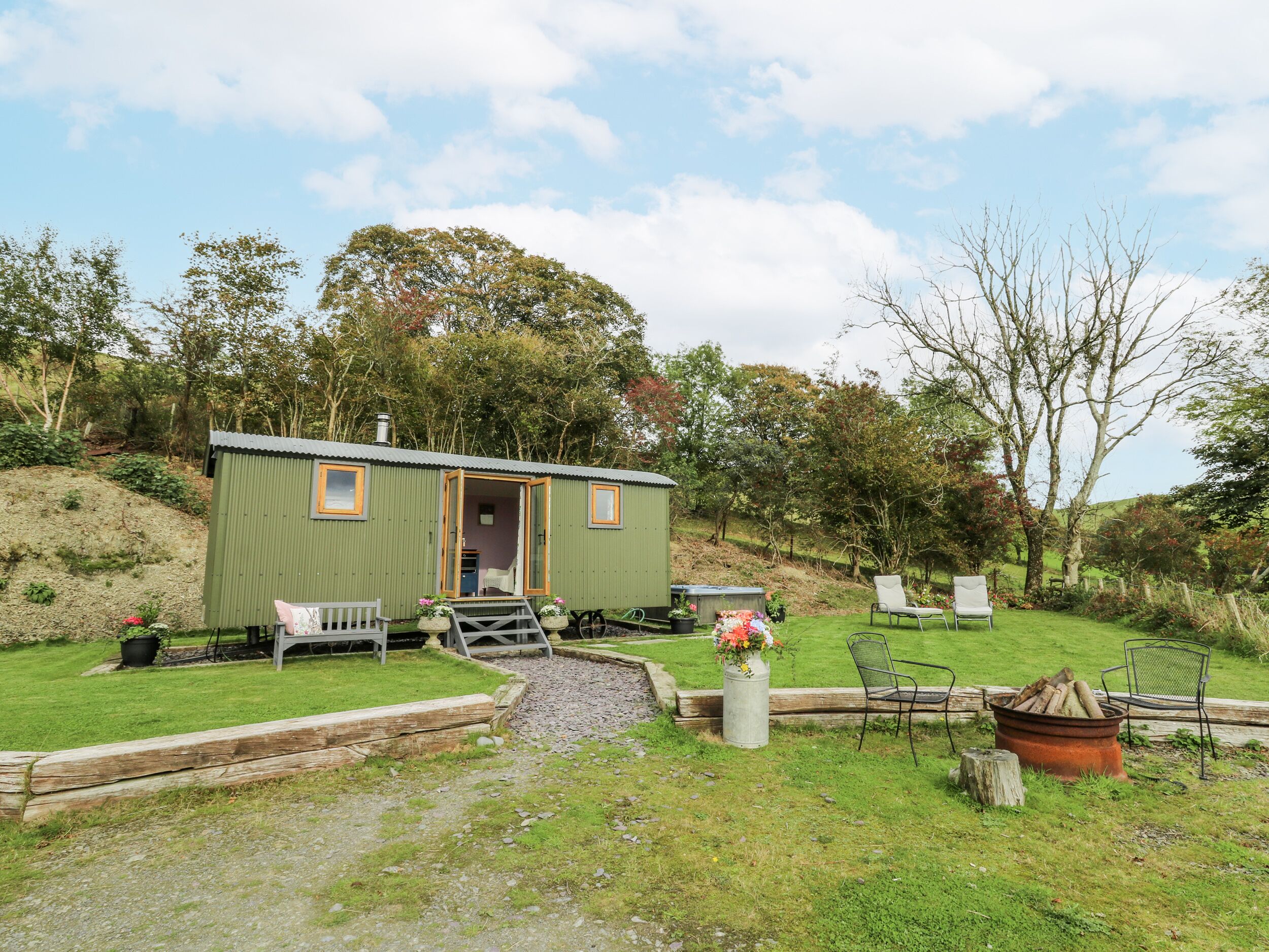 A small, green mobile home with a porch, a garden area with a fire pit, and outdoor seating.