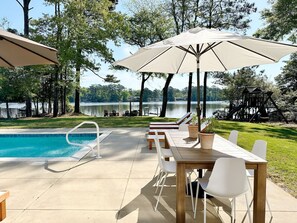 A poolside area with a wooden table and chairs, a pool, and umbrellas. - Bayfront With Dock Pool & Hot Tub! (Kitty Hawk)