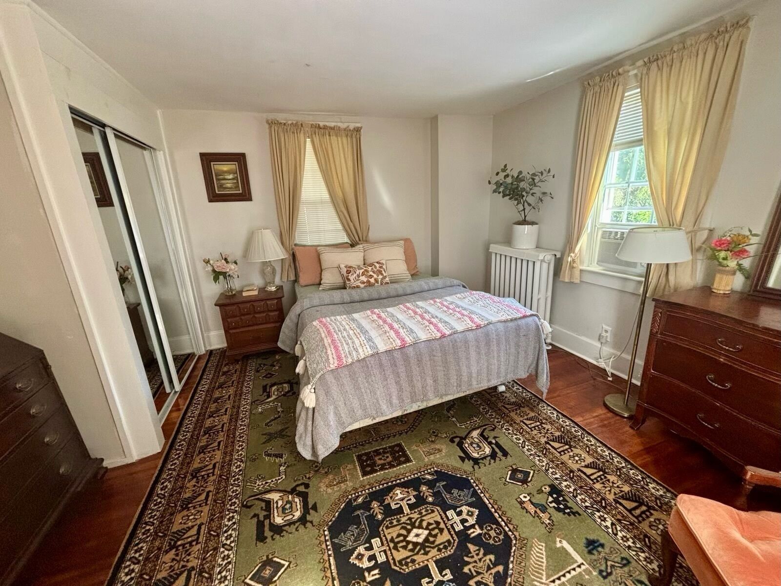 Een slaapkamer met een bed, een kaptafel, een nachtkastje, een radiator en een raam met gordijnen.
