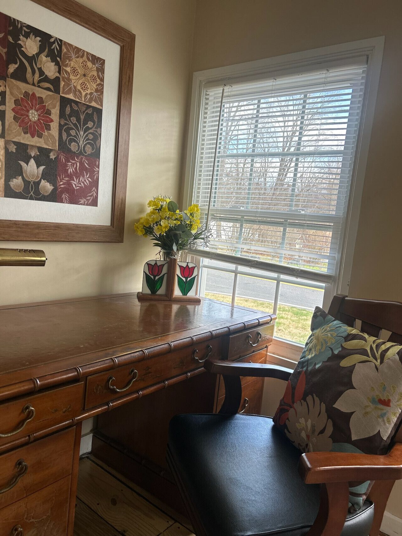 Een houten bureau met lades, een stoel, een lijst met bloemwerk en een vaas met gele bloemen op een vensterbank.