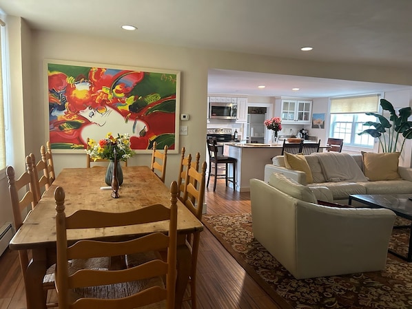 A dining area with a wooden table and chairs, a large colorful painting on the wall, and a kitchen in the background.