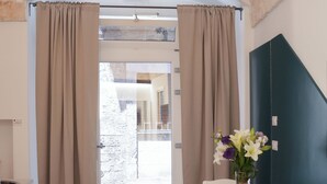 A room with a stone wall, a white door with beige curtains, a small table with a vase of flowers, and a white chair. - Giusy House (Ostuni)