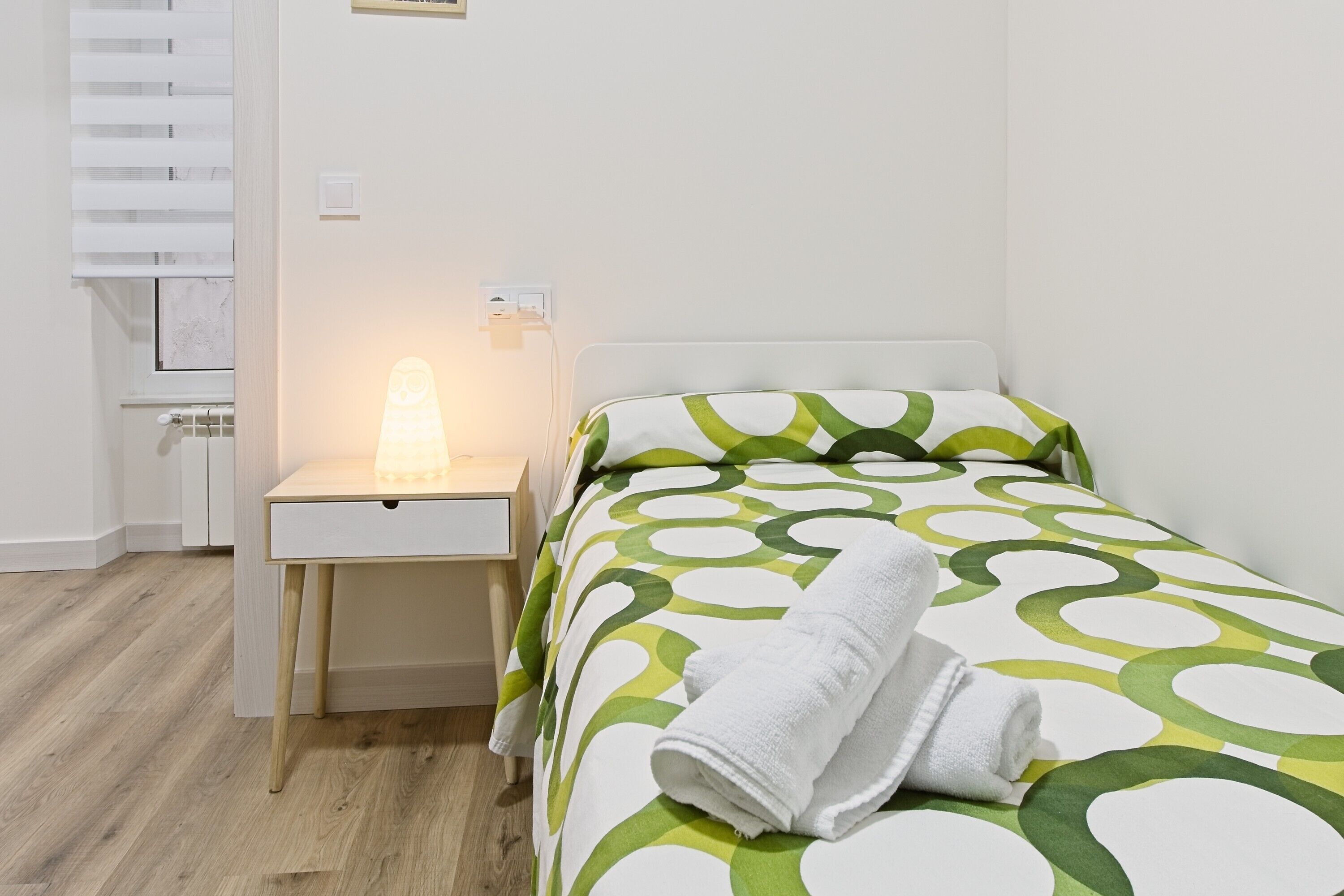 Ein Einzelbett mit einer grün-weißen Bettdecke, ein Handtuch auf dem Bett, ein Nachttisch mit Lampe und ein weißer Kleiderschrank.