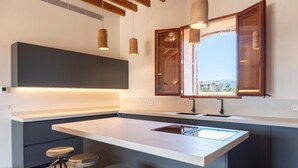 A modern kitchen with a large island, bar stools, pendant lighting, and a view through a window. - S' Hort de ca'n salvà (Santa Maria del Camí)