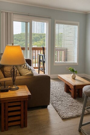 Una sala de estar con un sofá, una mesa con una lámpara, una silla y vistas a zonas verdes a través de amplios ventanales.