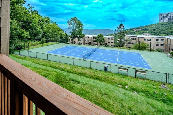 Una cancha de tenis con una terraza de madera al frente, rodeada de vegetación y edificios al fondo.