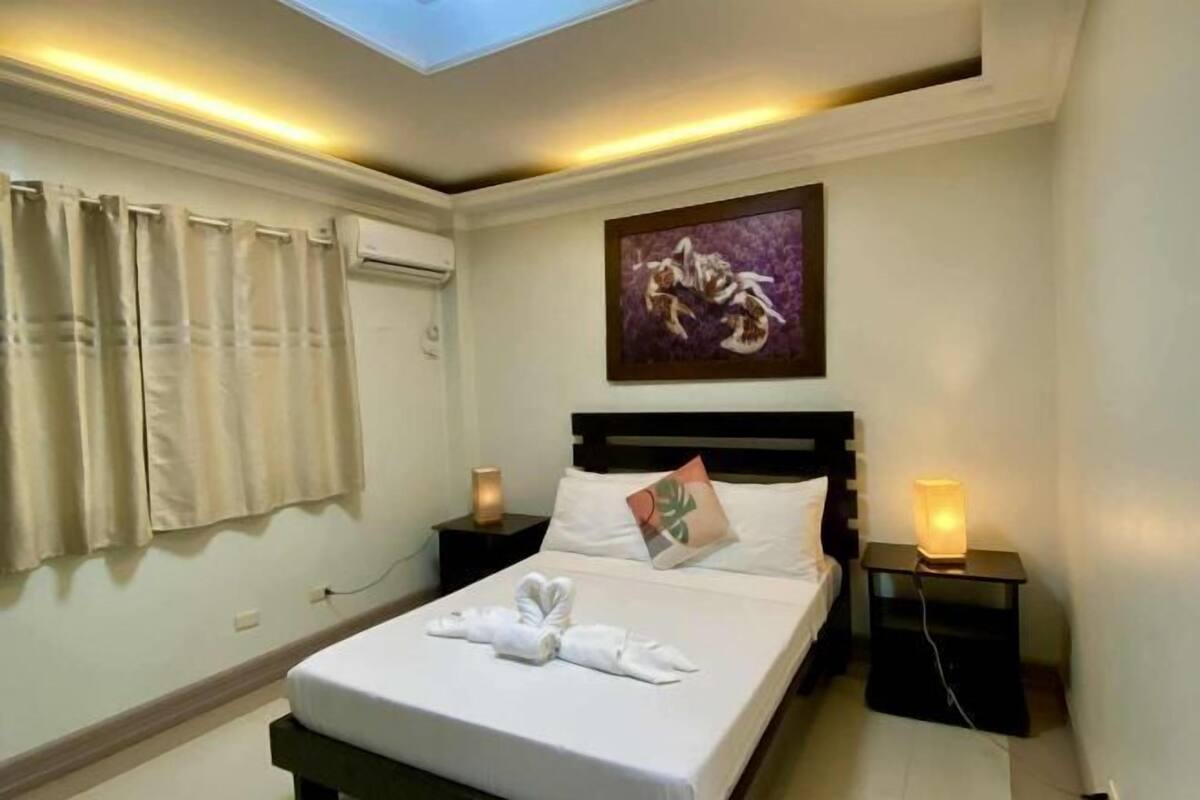 A hotel room with a bed, two bedside tables with lamps, a ceiling light, a window with curtains, and a wall-mounted artwork.