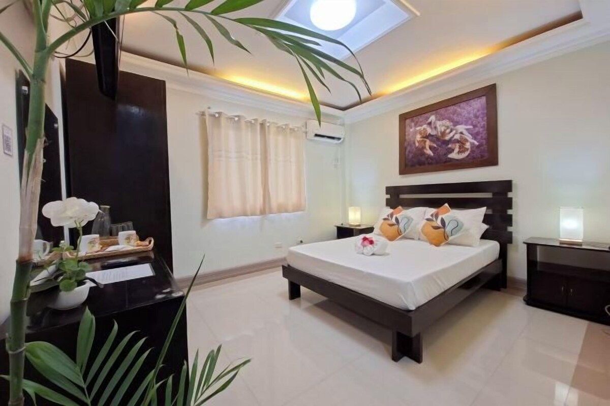 Habitación de hotel con cama, mesitas de noche, escritorio, planta en maceta, ventana con cortinas y un cuadro en la pared.