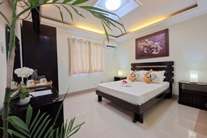 Habitación de hotel con cama, mesitas de noche, escritorio, planta en maceta, ventana con cortinas y un cuadro en la pared.
