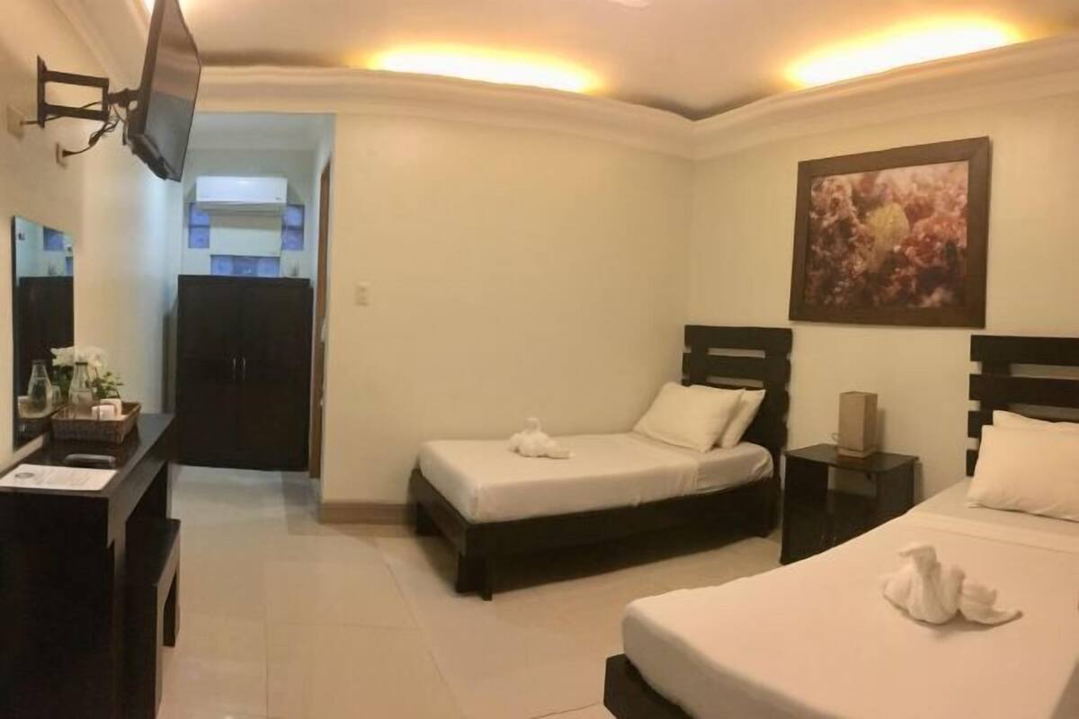 A hotel room with two beds, a desk with a mirror, a TV mounted on the wall, and a painting above the beds.