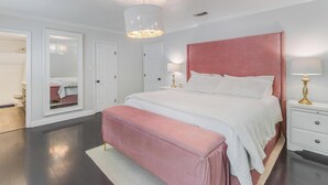 Un dormitorio con una cama grande, un cabecero rosa, una mesita de noche con una lámpara y un espejo en la pared.