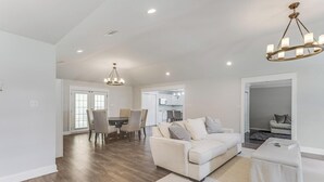 Amplia sala de estar con un sofá blanco, un comedor con mesa y sillas, y una cocina al fondo.