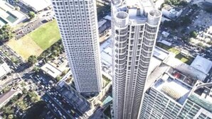 Un edificio alto con varios pisos, rodeado de otros edificios y una zona verde.