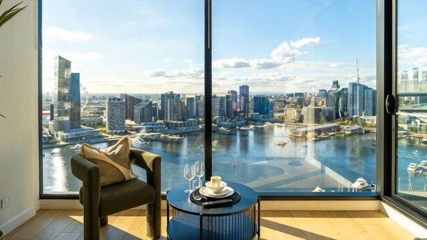 Habitación con una ventana grande que da a un paisaje urbano y al agua, una silla y una mesa con una copa de vino.