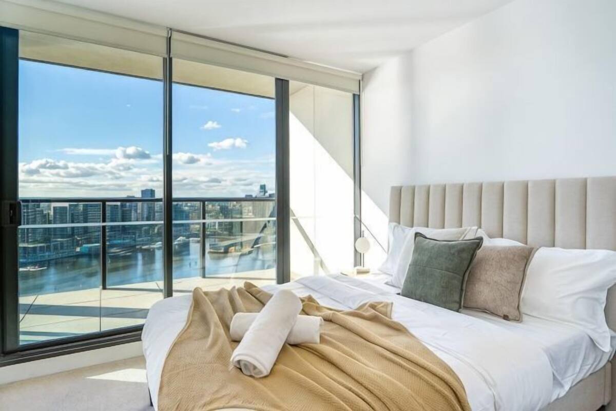 Una habitación de hotel moderna con una cama grande, vistas de la ciudad a través de ventanales del suelo al techo y un balcón.