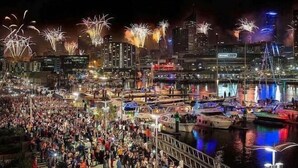Un paisaje urbano frente al mar con una gran multitud reunida cerca del agua, barcos amarrados y fuegos artificiales al fondo.