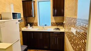 A compact kitchen with dark wooden cabinets, a white countertop, a microwave, a sink, and a window above it. - ALMOHAJER SERVICED APARTMENTS (Jeddah)