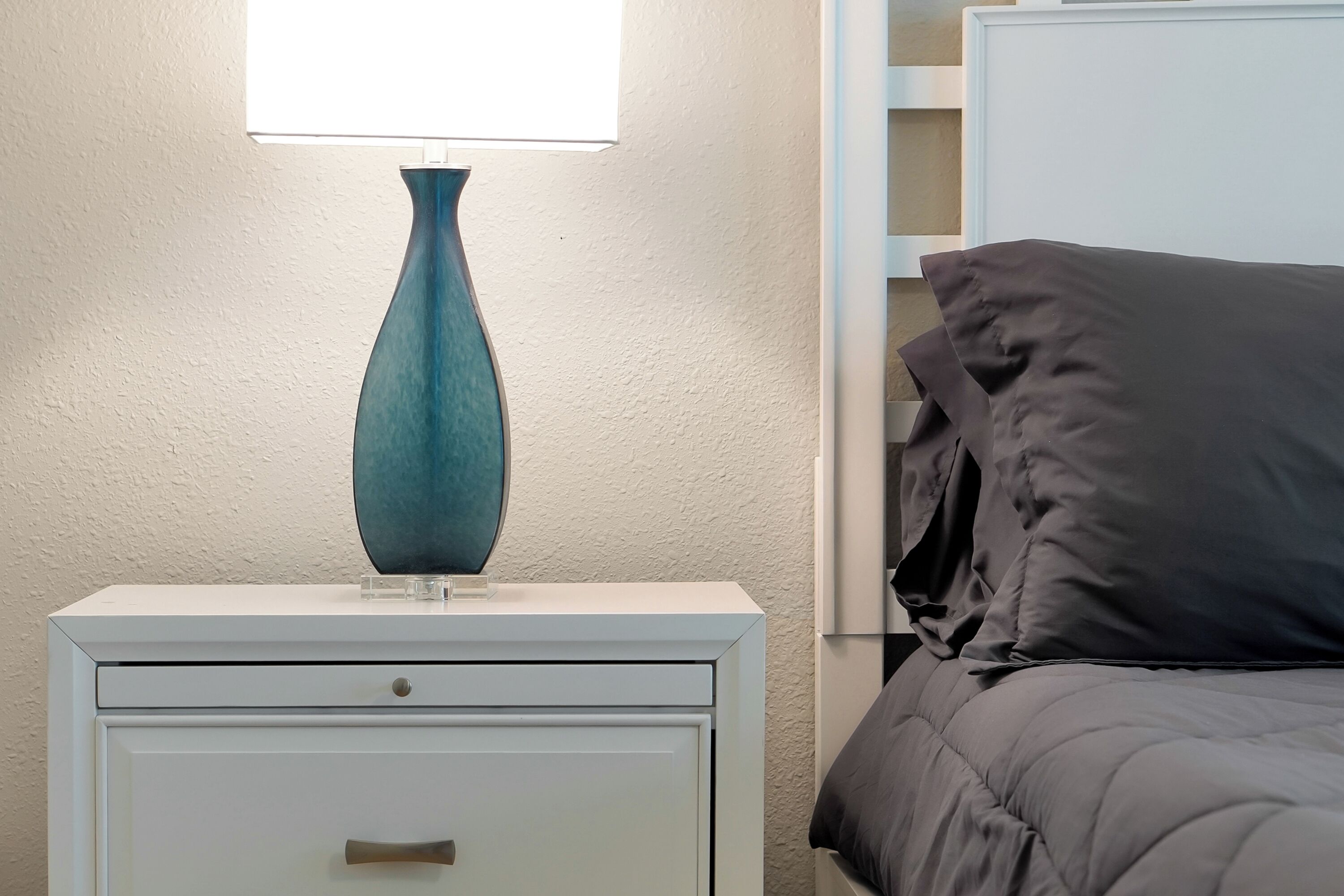 A white bedside table with a teal vase and a lamp, a gray bed with a dark gray pillow, and a white wall with a textured finish.