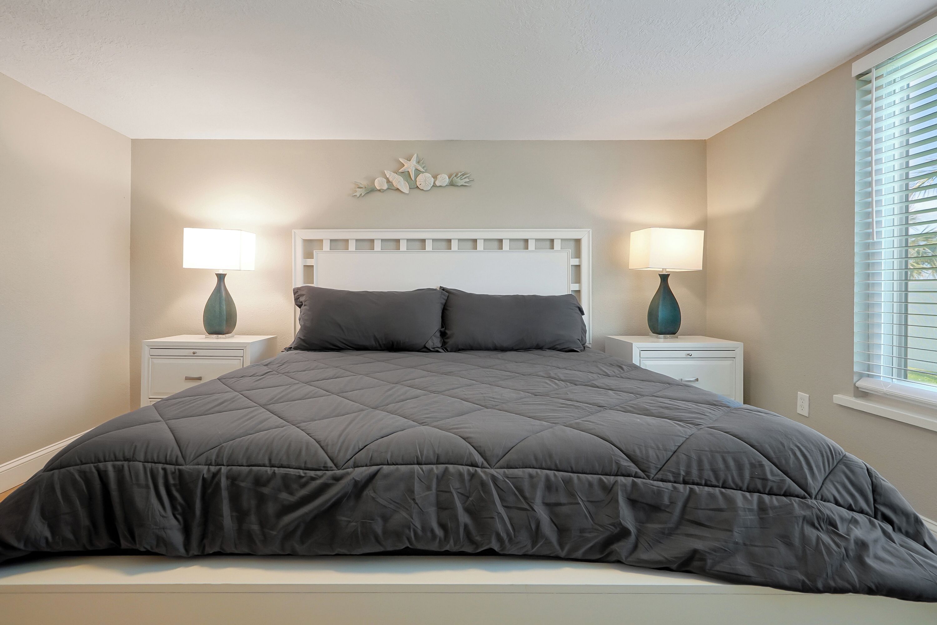A bedroom with a large bed, two bedside tables with lamps, a decorative wall piece above the bed, and a window with blinds.